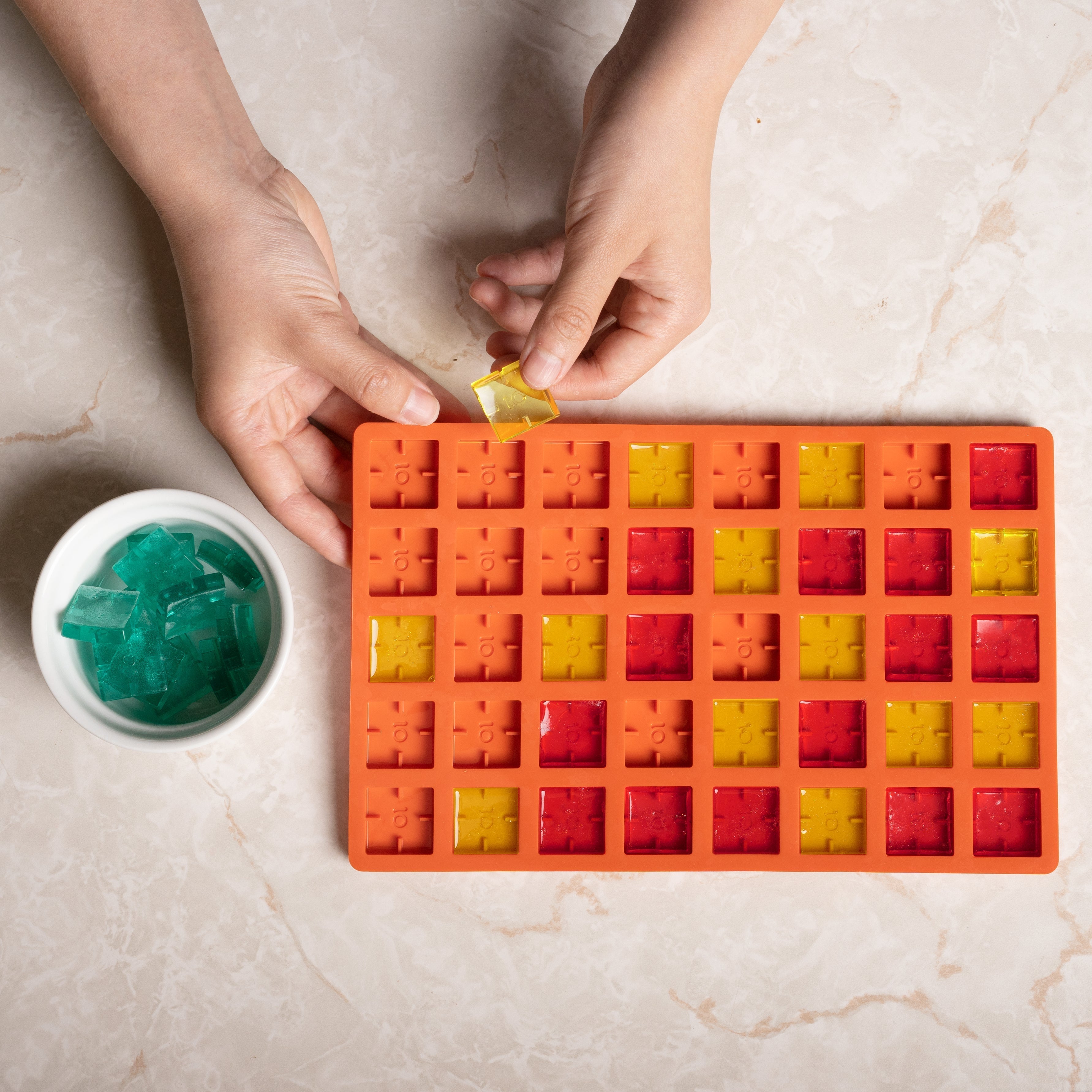 Person filling a colorful silicone ice cube tray with gummy bears on a marble surface.